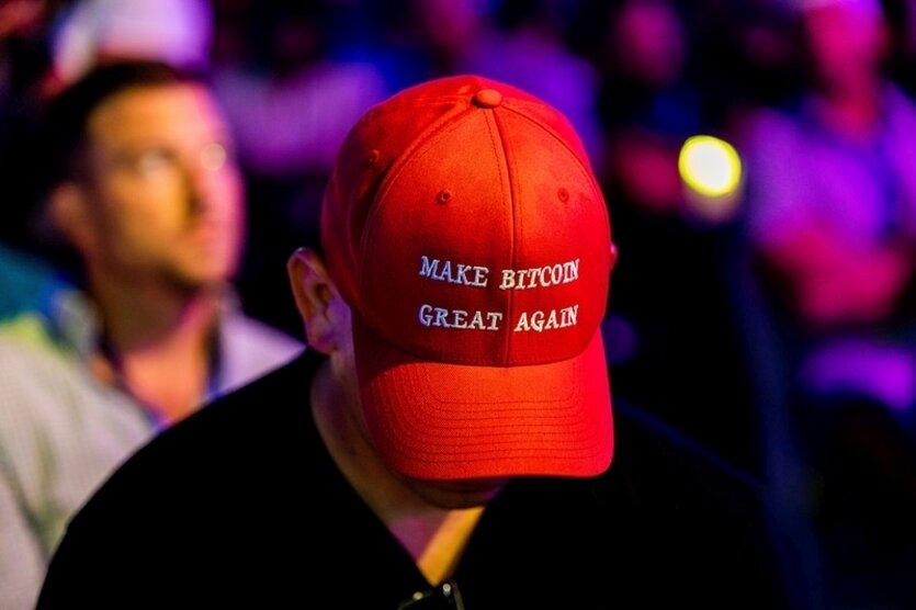 Eine Person mit schwarzem T-Shirt hält den Kopf gesenkt. Sie trägt eine rote Kappe, auf der "Make Bitcoin great again" steht. Im Hintergrund sitzen mehrere Menschen in lila Licht.