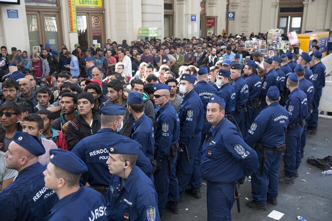 Eine Gruppe von Geflüchteten wartet Anfang September 2015 am Budapester Bahnhof Keleti auf die Weiterreise nach Österreich und Deutschland. Polizeibeamte stehen in einer Reihe direkt neben ihnen.