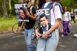 Zwei Menschen demonstrieren für die Freilassung von Osman Kavala und einer anderen in der Türkei Inhaftierten. Sie tragen Plakate mit den Koterfeis der beiden.