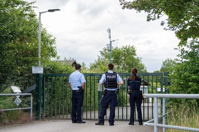 Zwei Polizisten und eine Polizistin stehen vor dem Eingang zu einer Kleingartenanlage