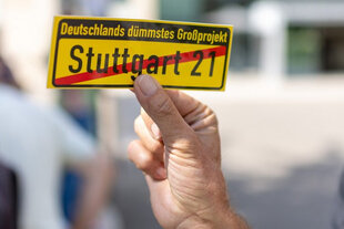 Eine Hand hält vor einer sonnenbeschienen Straße einen kleinen gelben Zettel hoch, der einer aufgehobenen Ortstafel nachempfunden ist und auf dem steht: „Deutschlands dümmstes Großprojekt – Stuttgart 21“.
