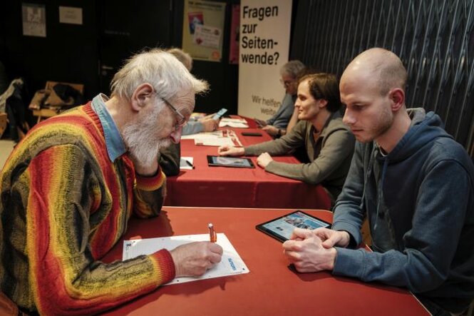 Es sind zwei Personen zu sehen, die sich unterhalten. Im Hintergrund steht ein Aufsteller, auf dem "Fragen zur Seitenwende?" geschrieben steht.
