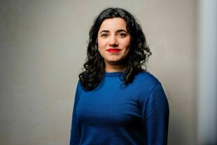 Misbah Khan steht in einem blauen Pullover vor einer grauen Wand