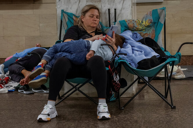 Eine Frau sitzt auf einem Campingstuhl in einm U-Bahnschacht. Sie hält ein schlafendes Kind auf dem Schoß