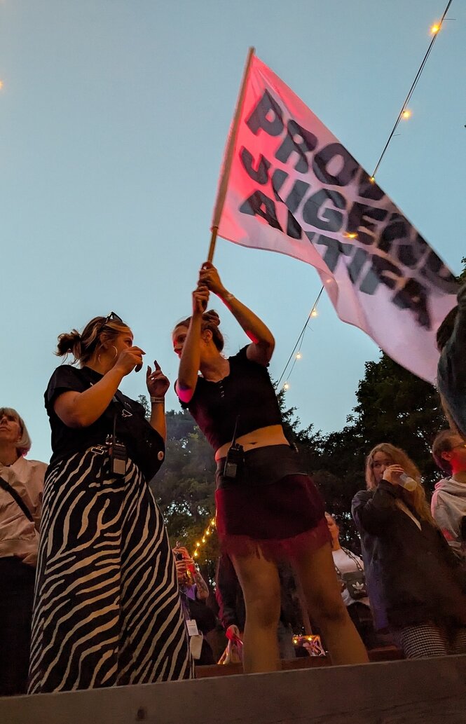 Zwei Frauen stehen nebeneinander in einer Menschenmenge, eine schwenkt eine Fahne mit dem Schriftzug "Provinz Jugend Antifa"