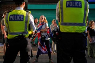 Eine Frau steht zwei Polizisten gegenüber, sie verschränkt ihre Arme und hat ein wütendes Gesicht. Über hren Schultern hängt die Fahne Großbritanniens