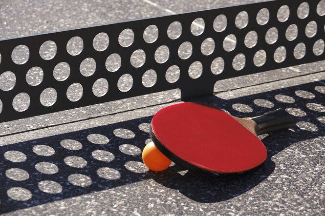 Ein Schläger und ein Ball liegen auf einer Steinplatte vor einem Gitter als Netz