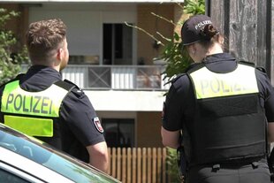 Zwei Polizist*innen stehen vor einem Haus, sie tragen Westen mit der Aufschrift Polizei