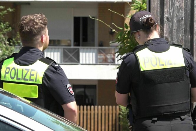 Zwei Polizist*innen stehen vor einem Haus, sie tragen Westen mit der Aufschrift Polizei