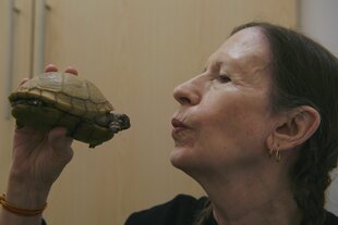 Sängerin Meredith Monk hält eine Schildkröte wie ein Mirkofon und deutet einen Kussmund an