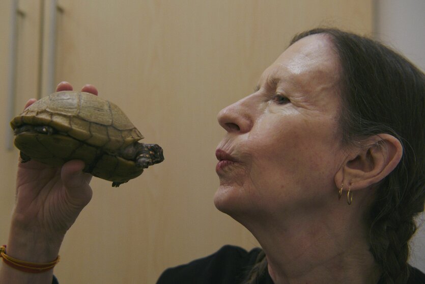 Sängerin Meredith Monk hält eine Schildkröte wie ein Mirkofon und deutet einen Kussmund an