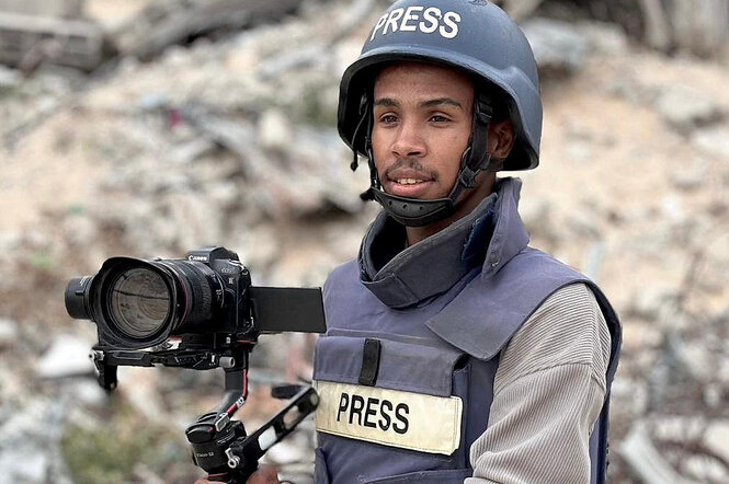 Mohammed Salama steht mit seiner Kamera vor Trümmern, er trägt einen Helm und Schutzweste mit der Aufschrift Presse