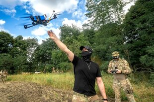Zwei vermummte Soldaten kontrollieren den Flug einer kleinen Drohne, einer hat das Konrollgerät in der Hand, der andere Mann reckt den Arm Richtung Drohne