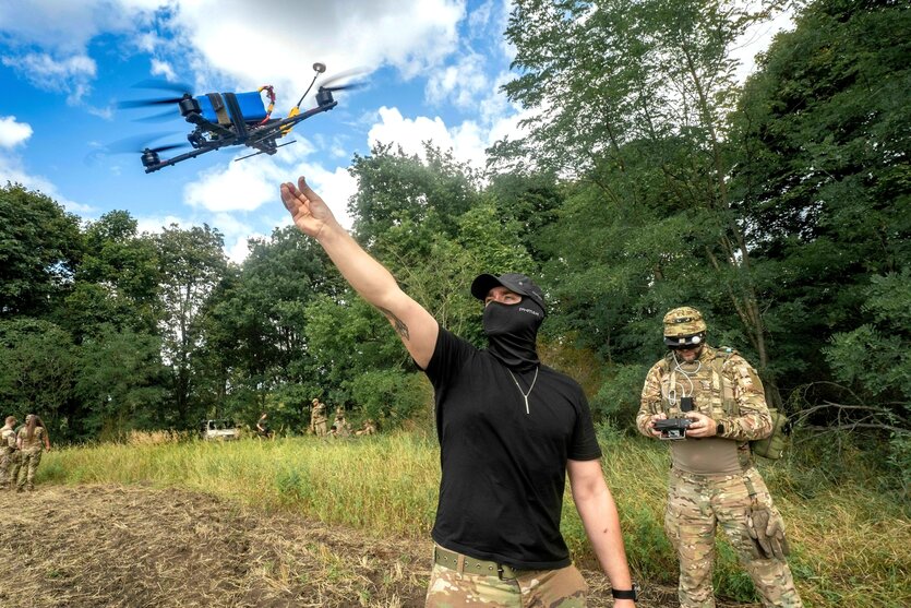 Zwei vermummte Soldaten kontrollieren den Flug einer kleinen Drohne, einer hat das Konrollgerät in der Hand, der andere Mann reckt den Arm Richtung Drohne