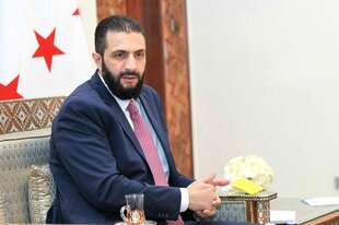 Ahmad al-Sharaa trägt Vollbart, Anzug und Krawatte. Er hat die Hände gefaltet und spricht. Er sitzt auf einem Sessel und hat ein goldenes Kissen im Rücken