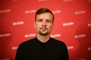 Porträt von Janis Ehling vor einer Wand mit dem Logo der Partei Die Linke
