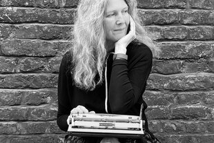 Schwarzweiß-Foto von Sabine Schiffner vor einer Backsteinmauer. Sie hat lange blonde Haare, auf ihrem Schoß ist eine Schreibmaschine und ihr Kinn stützt sie auf die linke Hand. Sie blickt nach links unten. Der Blick könnte als nachenklich beschrieben werden.