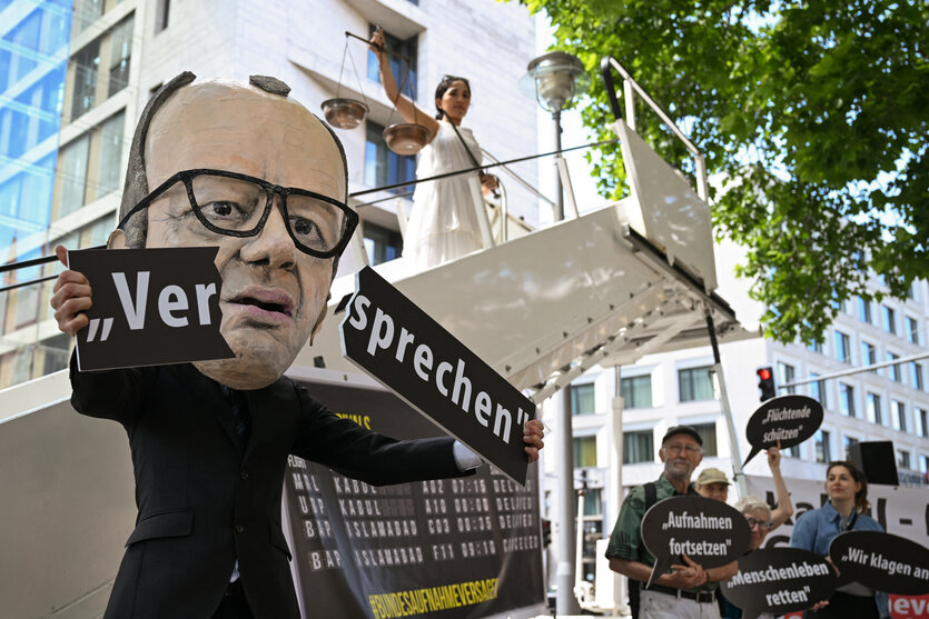 Demonstrierende protestieren gegen gebrochene Aufnahmezusagen der Bundesregierung. Ein Mensch trägt eine Maske von Kanzler Merz, und zerbricht symbolisch das Wort "Versprechen".