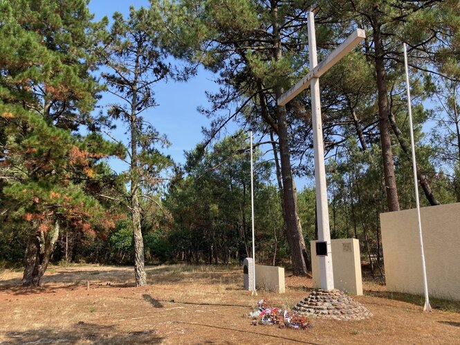 Eine Gedenkstätte im Kiefernwald, eine Kreuz mit einer Gedenktafel und einem Gedenkstein, vor dem Kränze abgelegt wurden