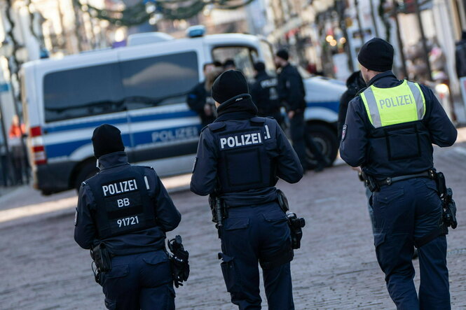 Drei Personen in Polizeiuniform von hinten gesehen, vor ihnen ein Gruppenfahrzeug