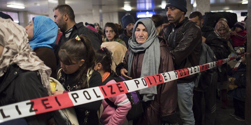 Mehrere Geflüchtete stehen hinter rot-weißem Flatterband am Flughafen Schönefeld in Berlin und warten darauf, dass es weiter geht.