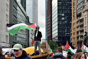 Zwischen den Hochhausschluchten Frankfurts zieht eine Pro-Palästina-Kundgebung, ein Kind, anscheinend auf den Schultern der Menge, hält eine Palästina-Flagge