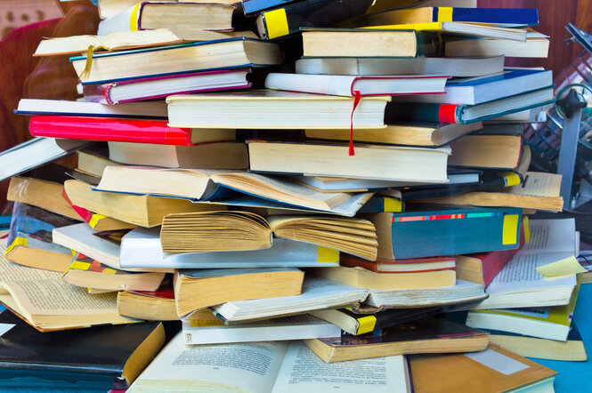 Ein großer, chaotischer Bücherstapel mit aufgeschlagenen und geschlossenen Büchern in verschiedenen Farben und Größen. Die Bücher sind nicht geordnet gestapelt, sondern liegen chaotisch aufeinander.