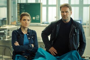 Eine Frau mit verschränkten Armen und ein Mann mit Händen in den Hüften stehen in einem gerichtsmedizinischen Raum.