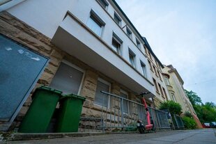 Dreigeschossige Immobilie vor teilweise bewölktem Himmel mit Mülltonnen und eScooter vor der Haustür.