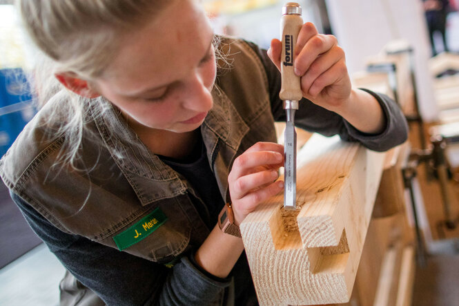 Eine weibliche Person in Handwerkskleidung bearbeitet in einer Tischlerei an ein Stück Holz.