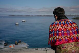 Frau in Parka, von hinten fotografiert am Meer sitzend