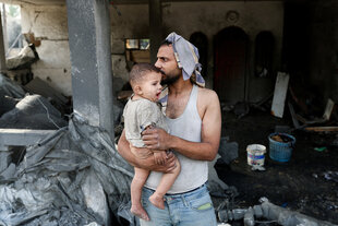 Ein Mann hält ein Kind in seinen Armen, er steht vor einer Hausruine im Gazastreifen