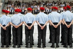 Soldat*innen stehen in einer Reihe vor Publkikum, sie tragen dunkle Hosen, hellblaue Hemden und ein rotes Barret