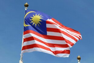 Die Flagge Malaysias weht vor blauem Hintergrund, rot-weisse Streifen und ein kleines blaues Quadrat oben links mit gelbem Mond und Sonne