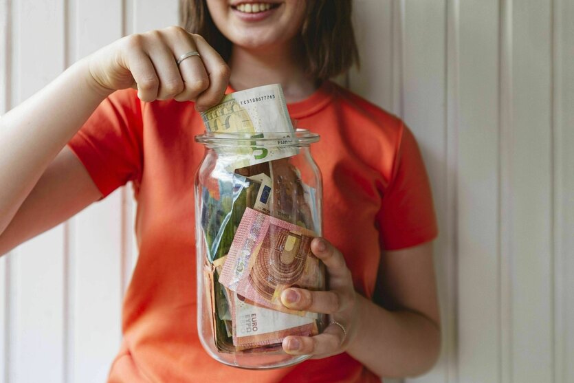 Junge Frau lächelt und steckt einen Fünf-Euro Schein in ein Glas mit vielen Geldscheinen
