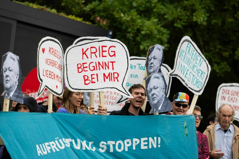 Protestaktion, Demonstrant*innen stehen hinder einem Banner mit der aufschrift: Aufrüstung stoppen, sie halten Schilder hoch wie : Krieg beginnt bei mir