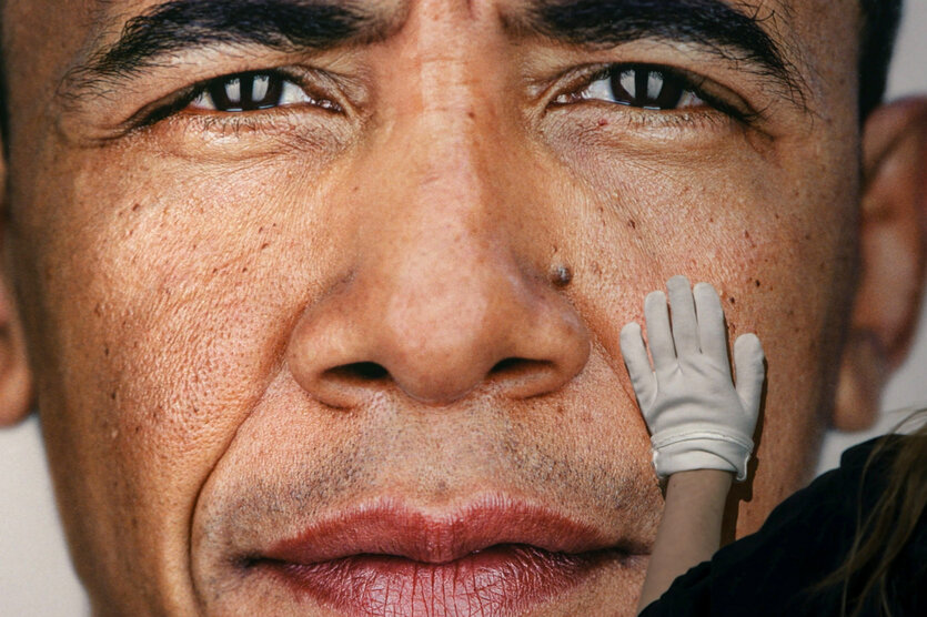 Nahaufnahme des Gesichts von Barack Obama mit einer weißen Hand in einem Handschuh, die die Wange berührt.