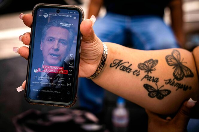 Eine Frau, mit Tätowierung auf dem Arm hält ein Handy in der Hand, auf dem das Porträt von Gavin Newsom zu sehen ist . Auf ihrem Arm steht: Nada es para siempre- (/Nichts ist für immer)