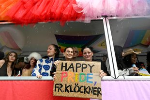 Teilnehmer des Christopher-Street-Day (CSD) Umzugs mit bunten Outfits au einem Wagen: Zwei Frauen halten ein Schild in der Hand: Happy Pride Frau Klöckner