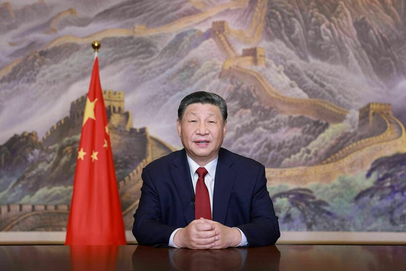 Xi Jinping sitzt vor einem gemalten Bild das die chinesische Mauer zeigt. Neben ihm hängt schlaff eine chinesische Fahne