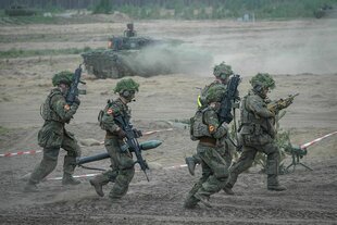 Fünf bewaffnete Soldaten in Tarnunfirom rennen über eine sandige Piste, im Hintergrund ist ein Panzer zu sehen, der Staub aufwirbelt