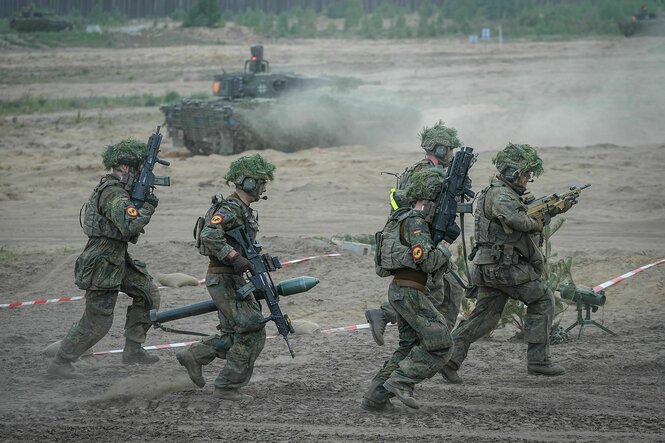 Fünf bewaffnete Soldaten in Tarnunfirom rennen über eine sandige Piste, im Hintergrund ist ein Panzer zu sehen, der Staub aufwirbelt