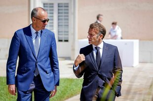 Friedrich Merz und Emmanuel Macron spazieren zusammen in der Sonne, Friedrich Merz trägt eine Sonnenbrille