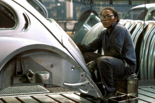 Ein Arbeiter mit Schutzbrille sitzt in einer Werkshalle vor einem VW-Käfer ohne Räder