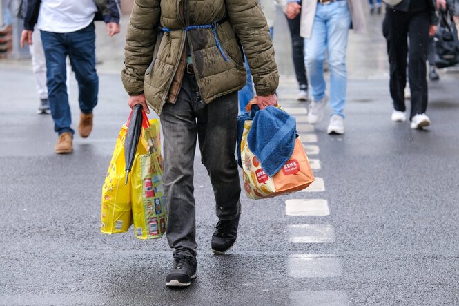 Straßenübergang, eine Person mit mehreren abgewetzten Einkaufstaschen läuft über die Straße, wir sehen vom Oberkörper abwärts. Die Jack ist alt, wirkt schäbbig und wird von einem Transportgummi zusammengehalten