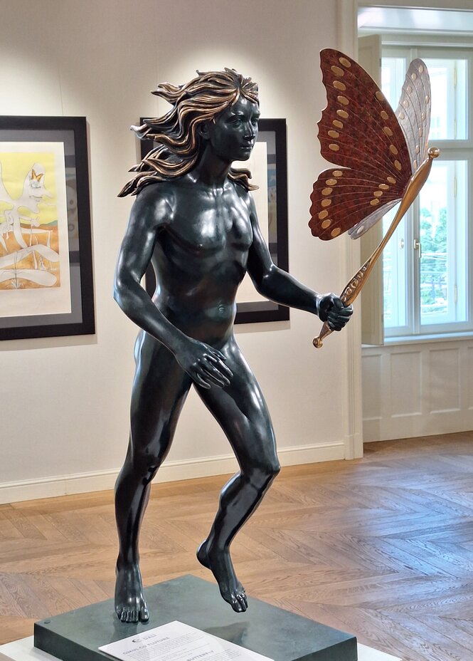 Auf einem flachen Sockel steht eine Skulptur von Dali, ein Mensch mit langen Haaren und großem Schmetterling, den er an einem Stab in der Hand hält