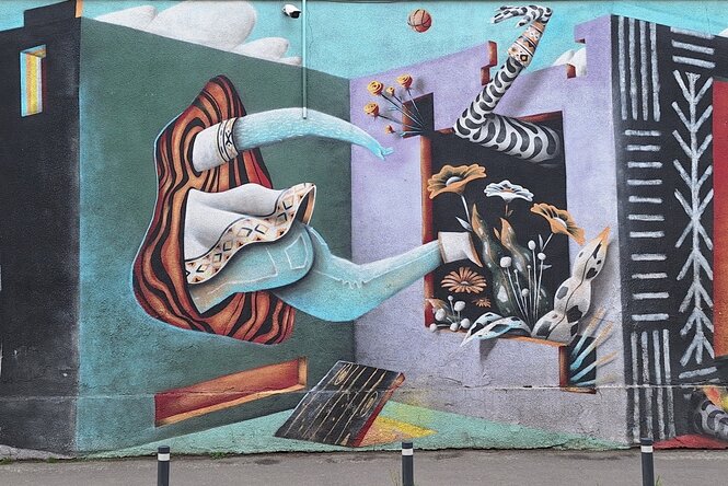 Auf eine Wand ist eine Ecke von Mauern gemalt. Einmal schaut ein Bein heraus, einmal Rock, Arm und Fuß.