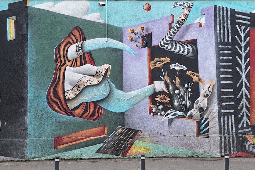 Auf eine Wand ist eine Ecke von Mauern gemalt. Einmal schaut ein Bein heraus, einmal Rock, Arm und Fuß.