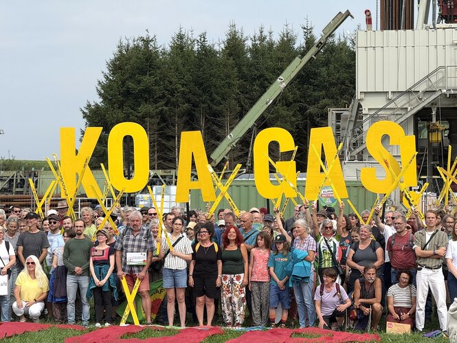 Protestaktion vor dem Bohrturm, die Demonstranten halten gelbe Kreuze hoch und tragen große gelbe Buchstaben mit der Info: KOA GAS !