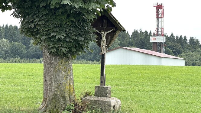 Ein Wegkreuz steht an einem Baum und im Hintergrund steht der Bohrturm, der untere Teil wird von einem Wellblechgebäude verdeckt